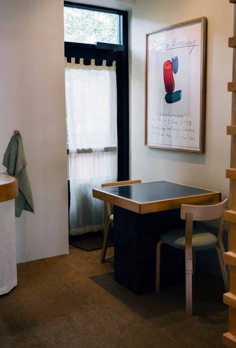 Small dining table with two chairs beside a wall, featuring a large framed art print and a window with sheer curtains.
