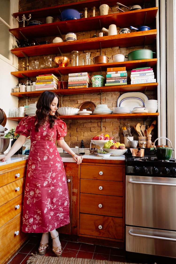 Woman in a red floral dress stands in a cozy kitchen with open wooden shelves.