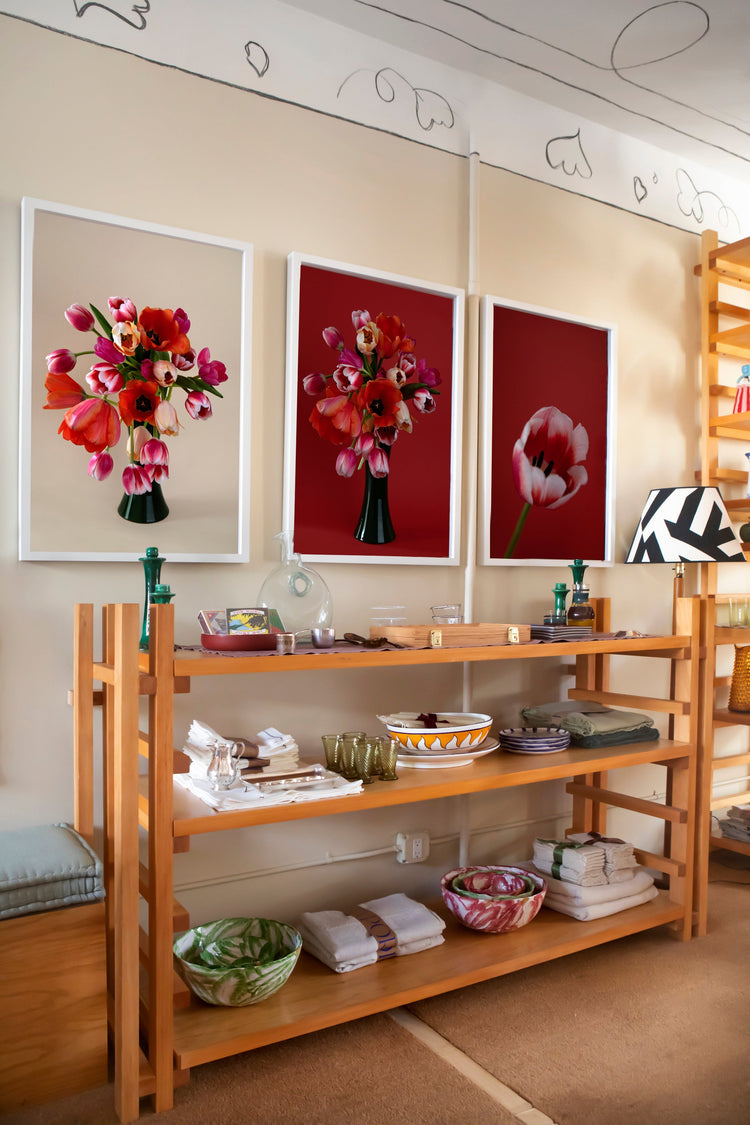 ChatGPT said: Wooden shelving unit with bowls, plates, and folded linens, set against a wall with three framed floral prints in red tones.