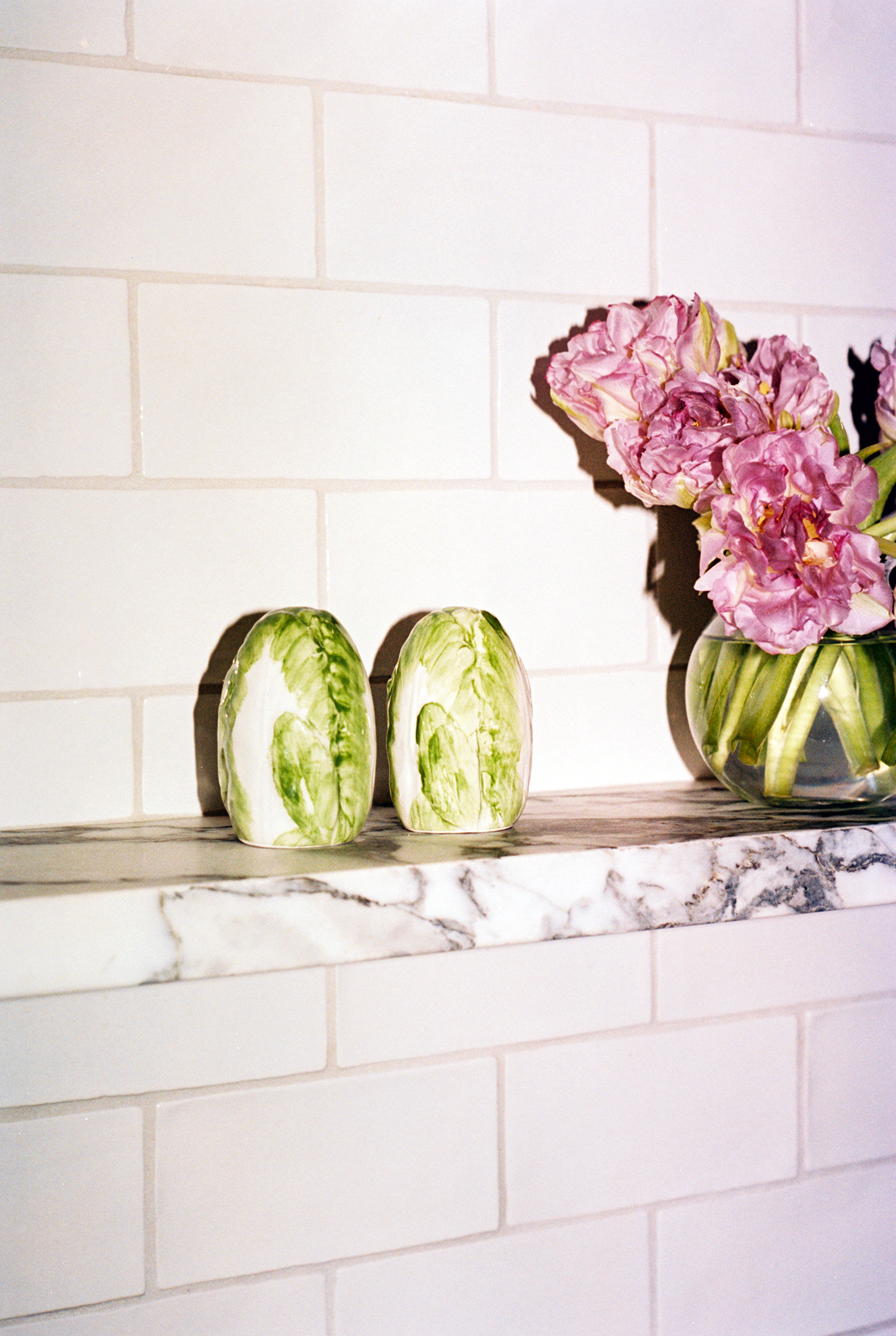 Two halves of a cabbage, a glass vase with pink flowers, and the Radicchio Salt and Pepper Shakers are displayed on a marble shelf against a tiled wall.