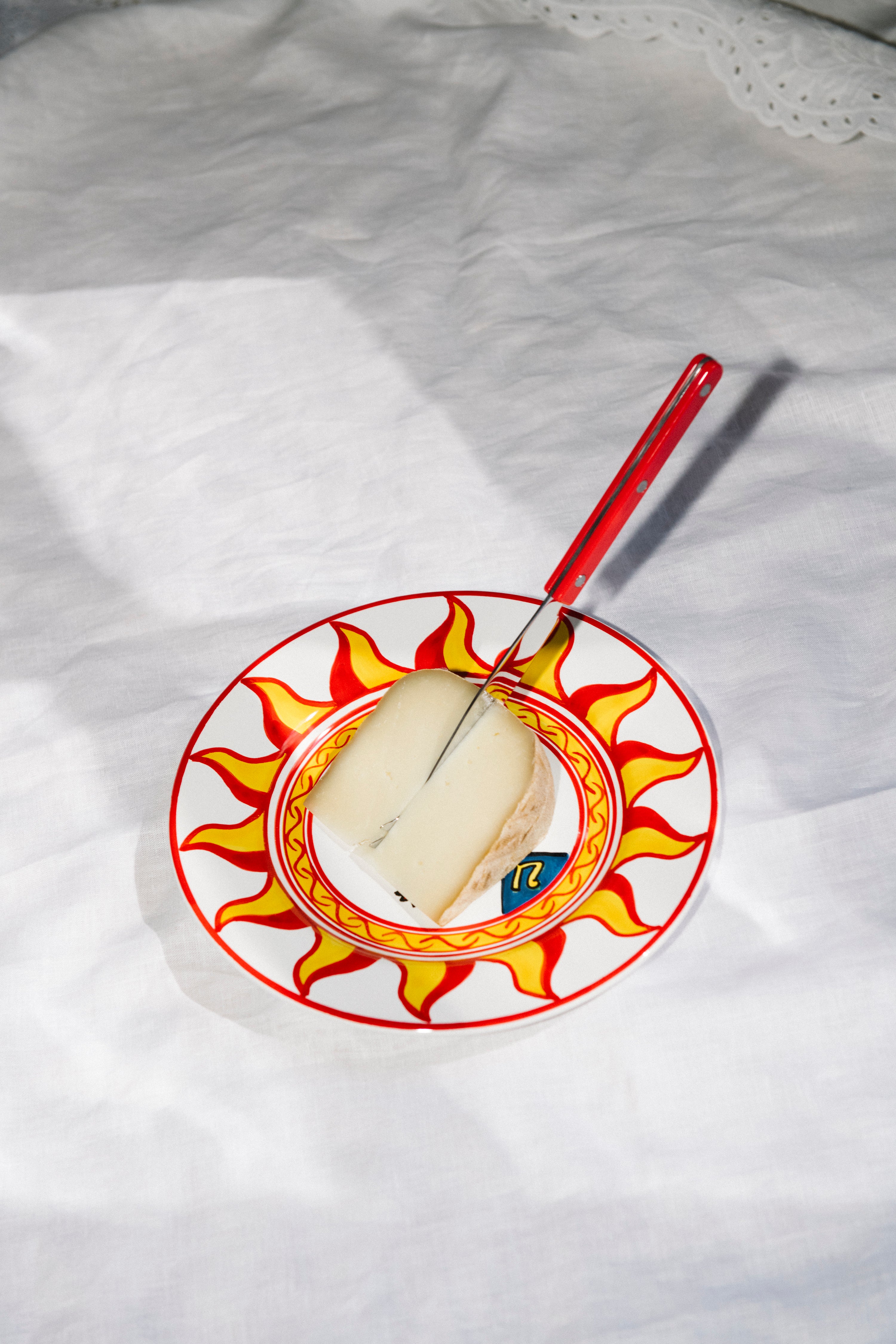 A large cheese knife with a red handle rests on a wedge of cheese atop a sun-patterned plate, all set against a white fabric background.