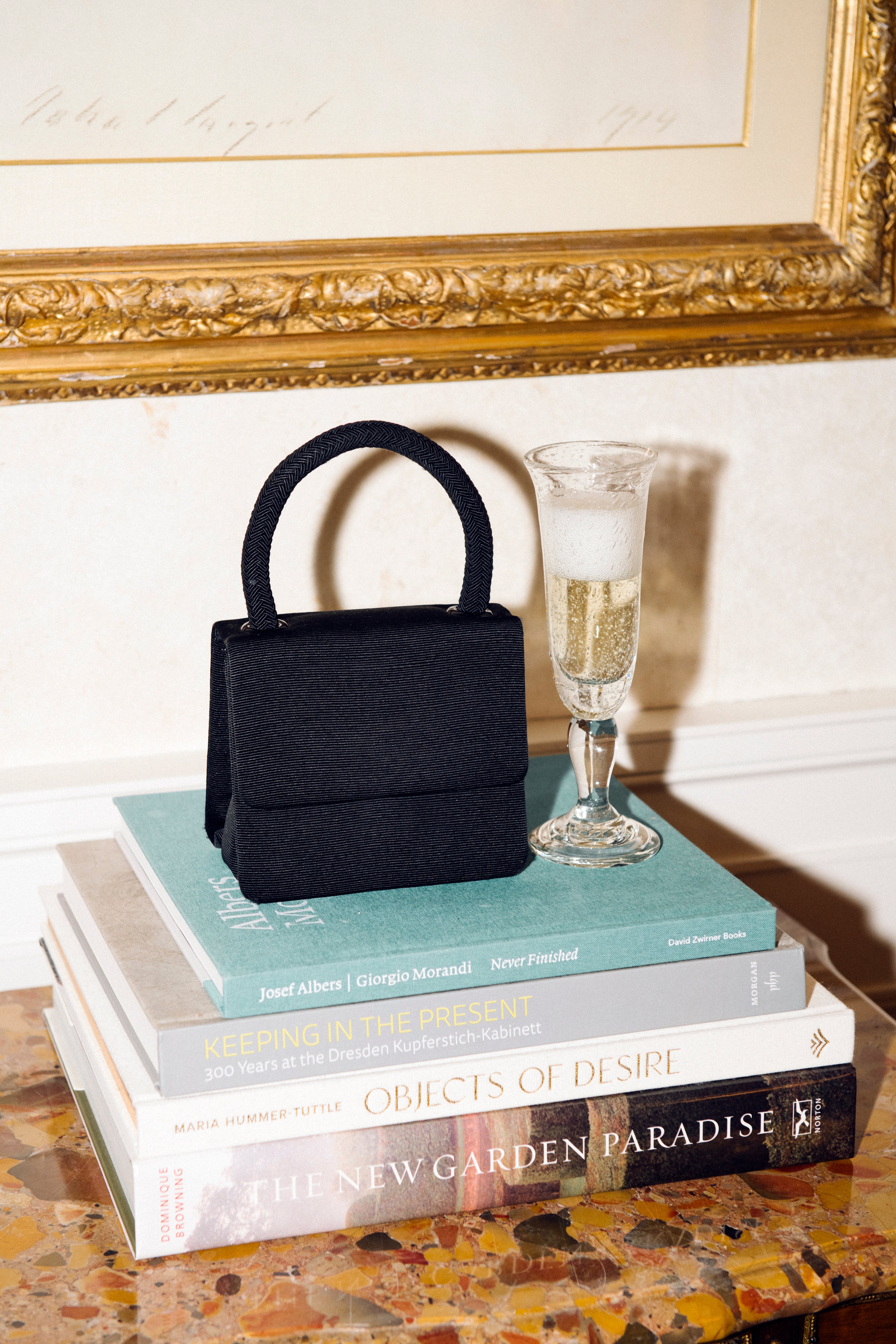 A Lauren Champagne Glass, filled with bubbly champagne, sits beside a small black handbag on a stack of books atop a marble surface, with framed artwork in the background.