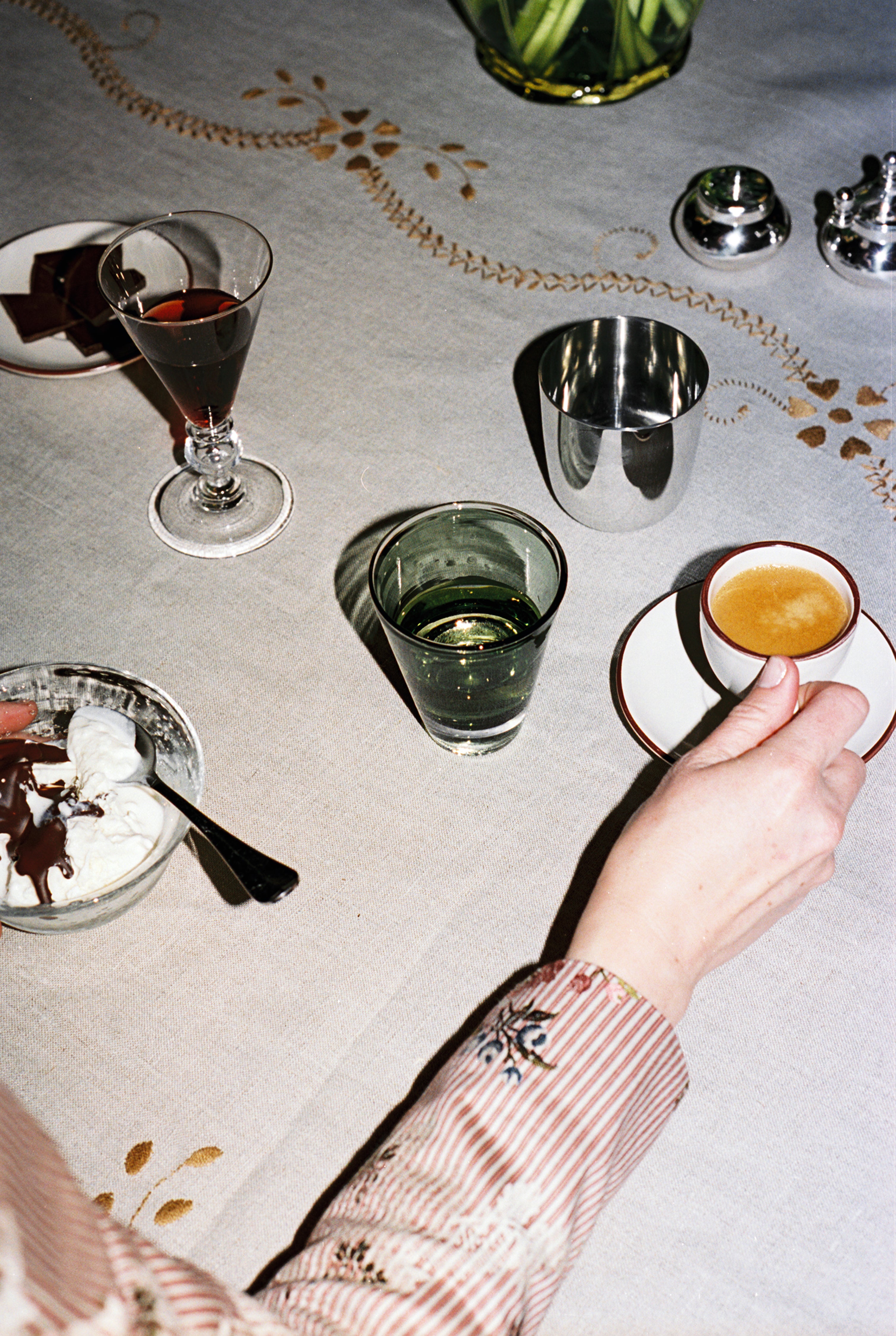 A person holds an espresso cup at a table featuring a Gem Glass, Set of 2, alongside a dessert bowl, glass of water, wine, and luxurious drinking glasses on a patterned tablecloth.