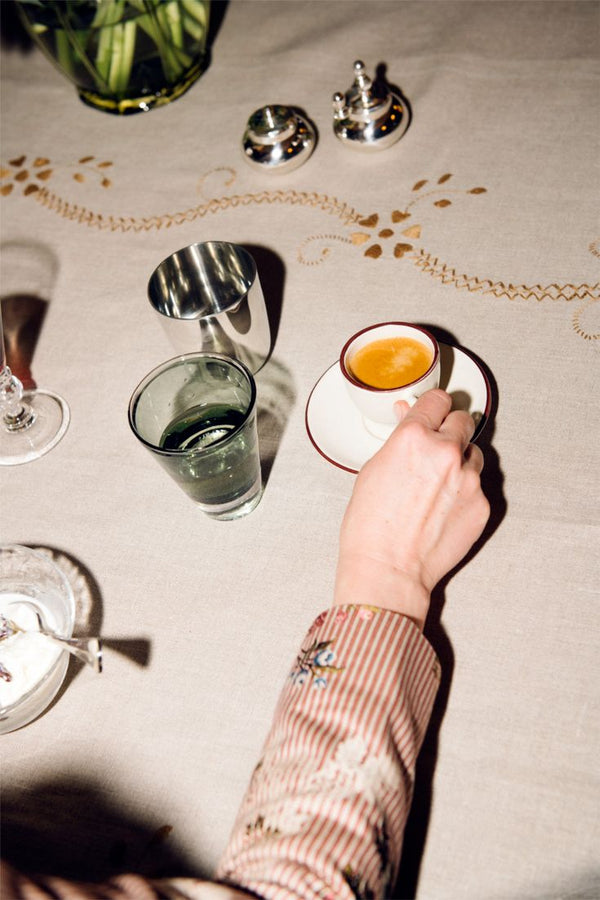 A person holding a Gio Espresso Set cup at a table set with a glass of water, coffee cup and saucer, silver containers, and an embroidered tablecloth.