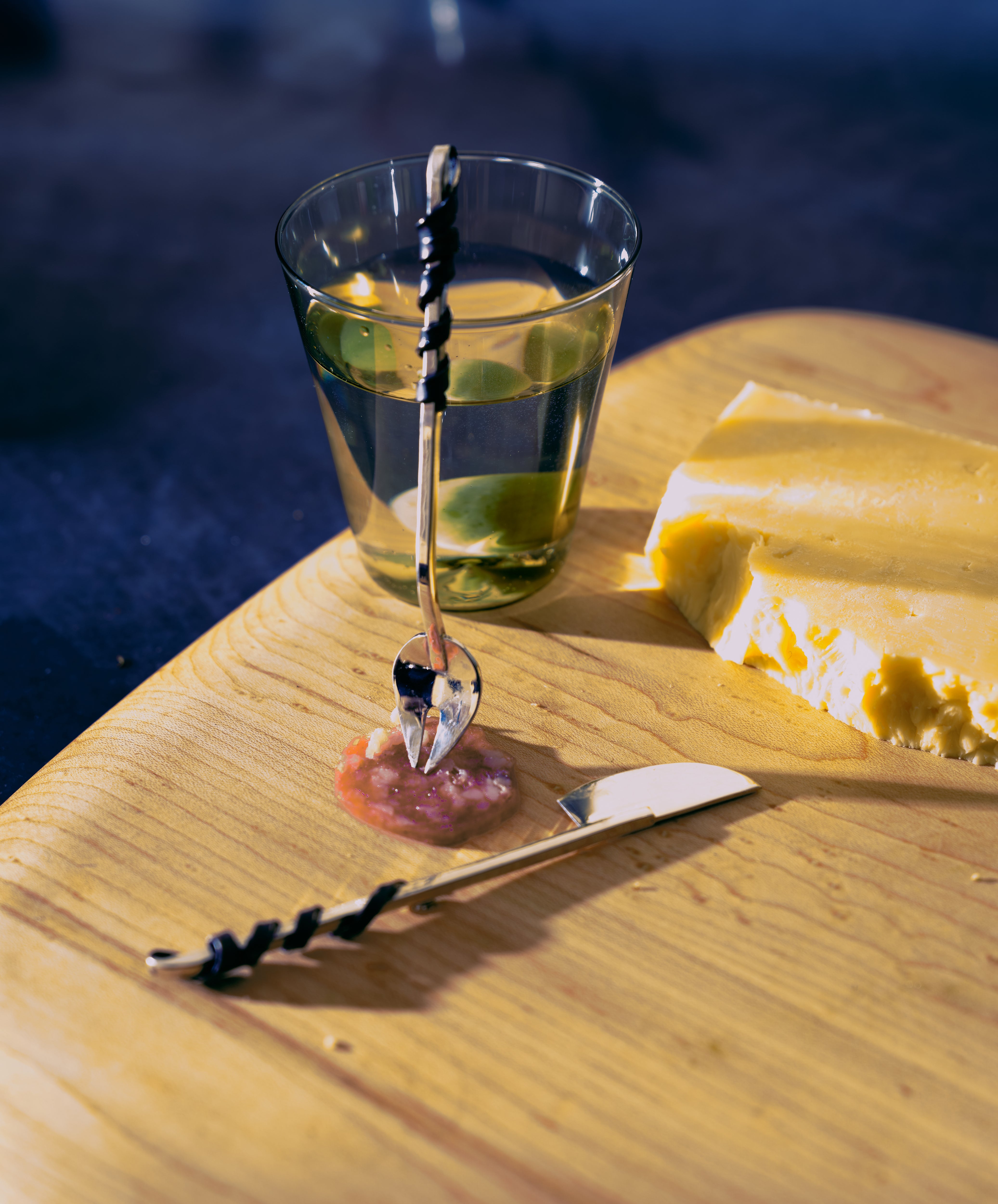 The Heath Wagoner | Aperitivo Set features a wooden cutting board adorned with water, yellow cheese, a cheese knife, and a black-handled spoon on minced condiments—true jewelry for your aperitivo table.