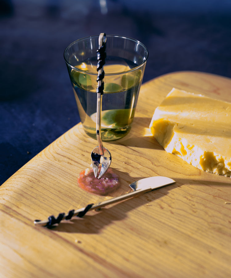 The Heath Wagoner | Aperitivo Set features a wooden cutting board adorned with water, yellow cheese, a cheese knife, and a black-handled spoon on minced condiments—true jewelry for your aperitivo table.