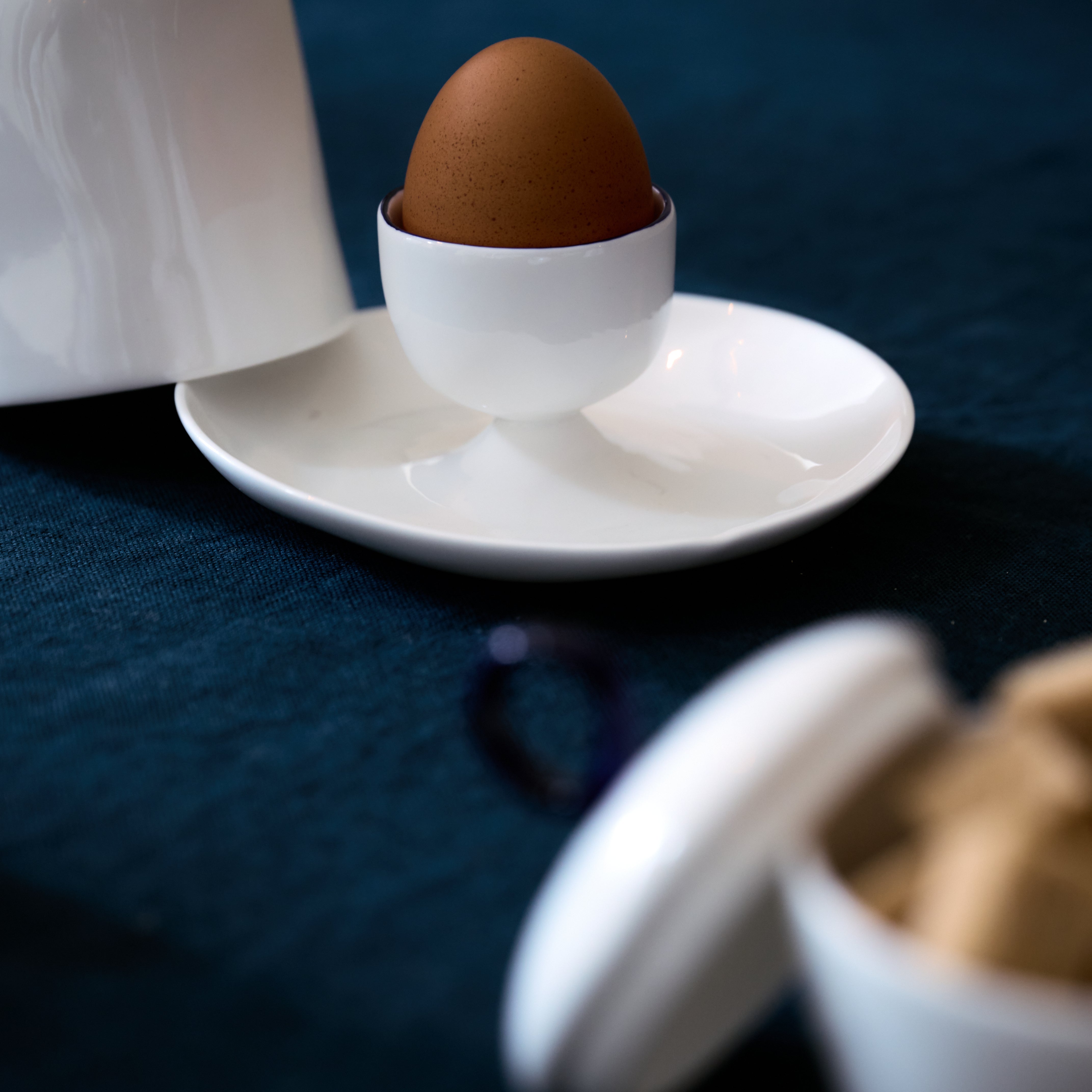 The Egg Cup and Lid holds a brown egg upright on a white plate, with a jar and container nearby on a dark blue tablecloth—ideal for your breakfast needs.