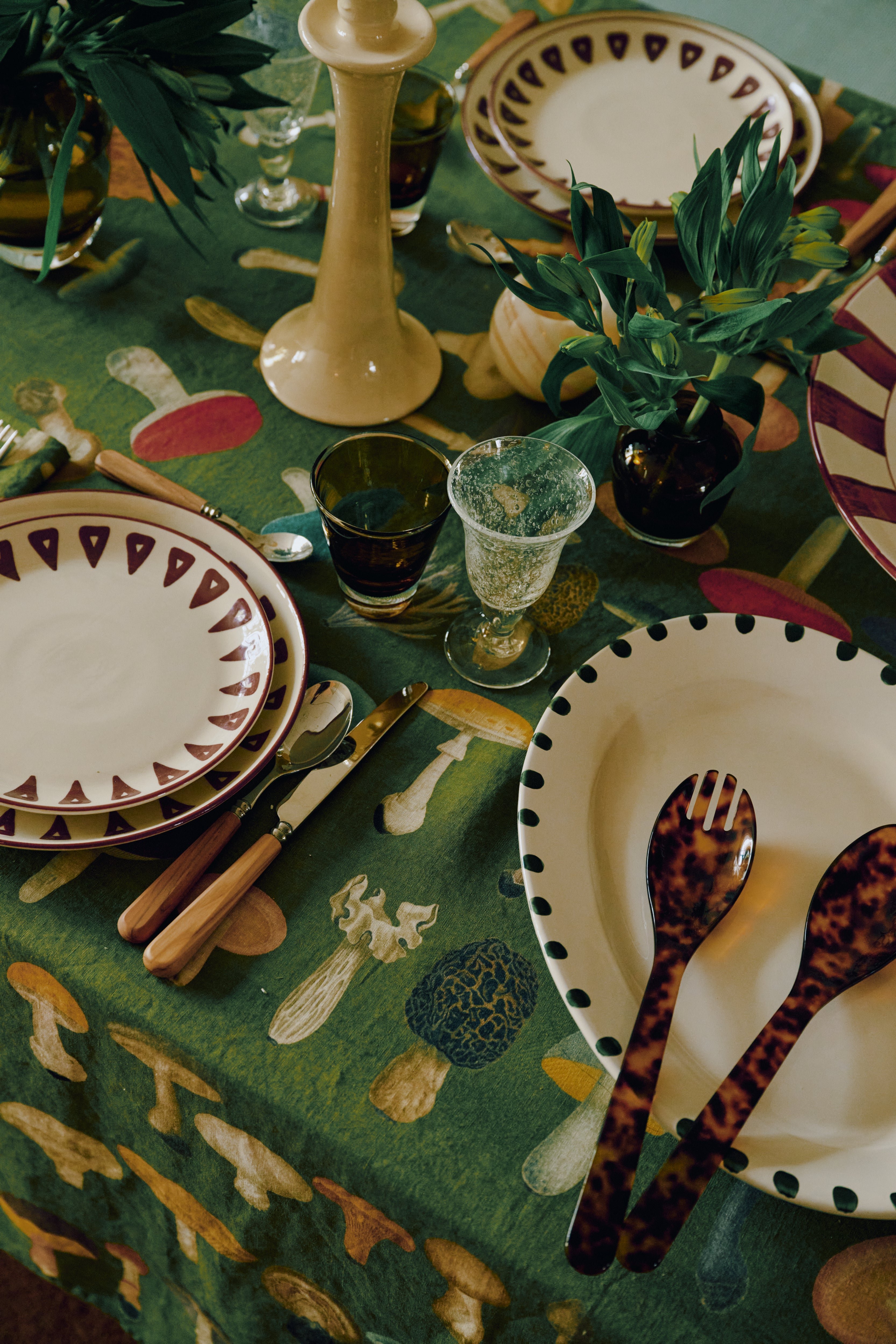 Table set with decorative plates, cutlery, green plants, and a green tablecloth featuring mushroom illustrations, complemented by the Gem Glass, Set of 2 luxury glassware.