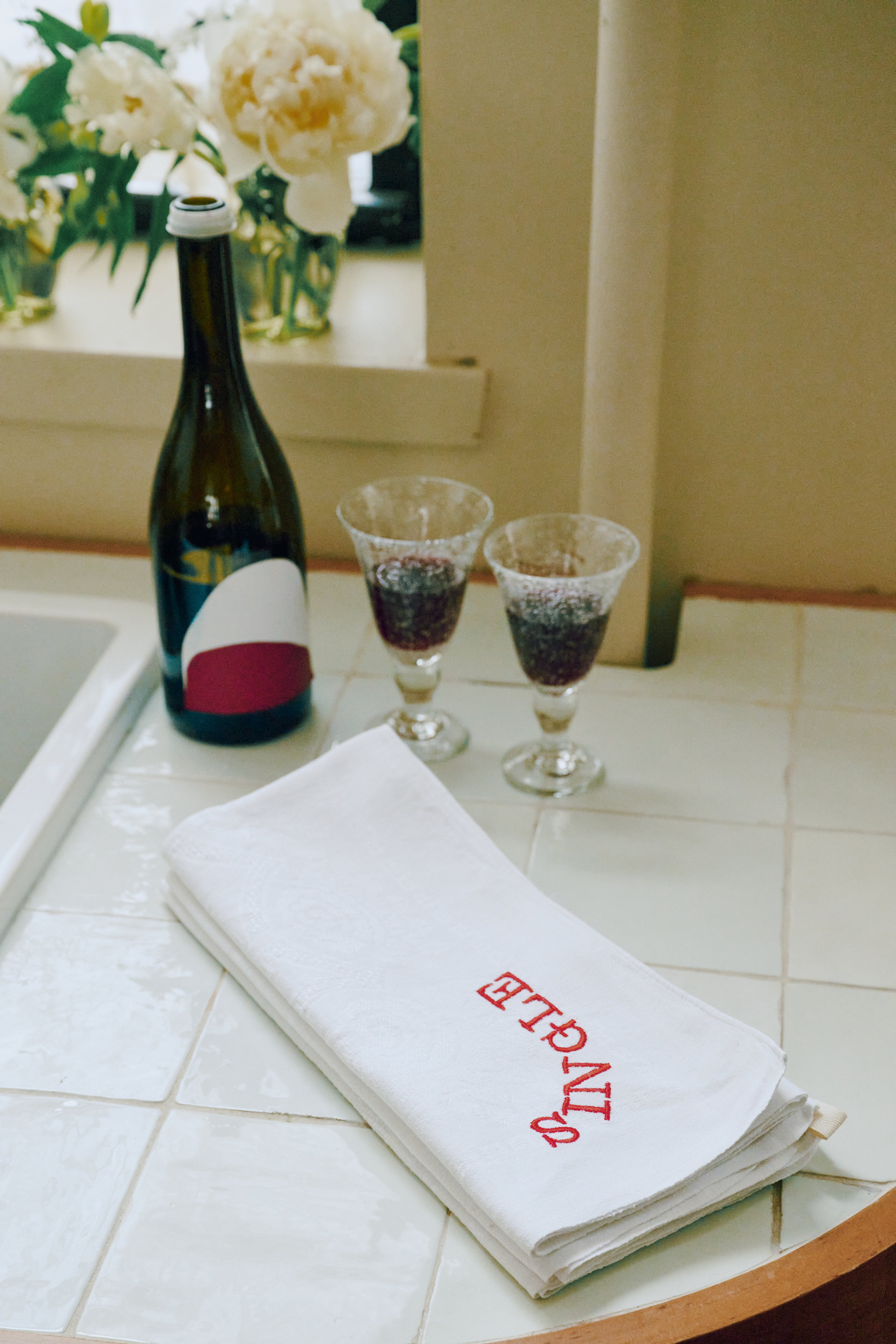A bottle of wine and two filled glasses sit on a tiled countertop beside the Relationship Status Napkin, Set of 6—one folded with ENTRE in red. Flowers on the windowsill evoke a fun dinner party mood.