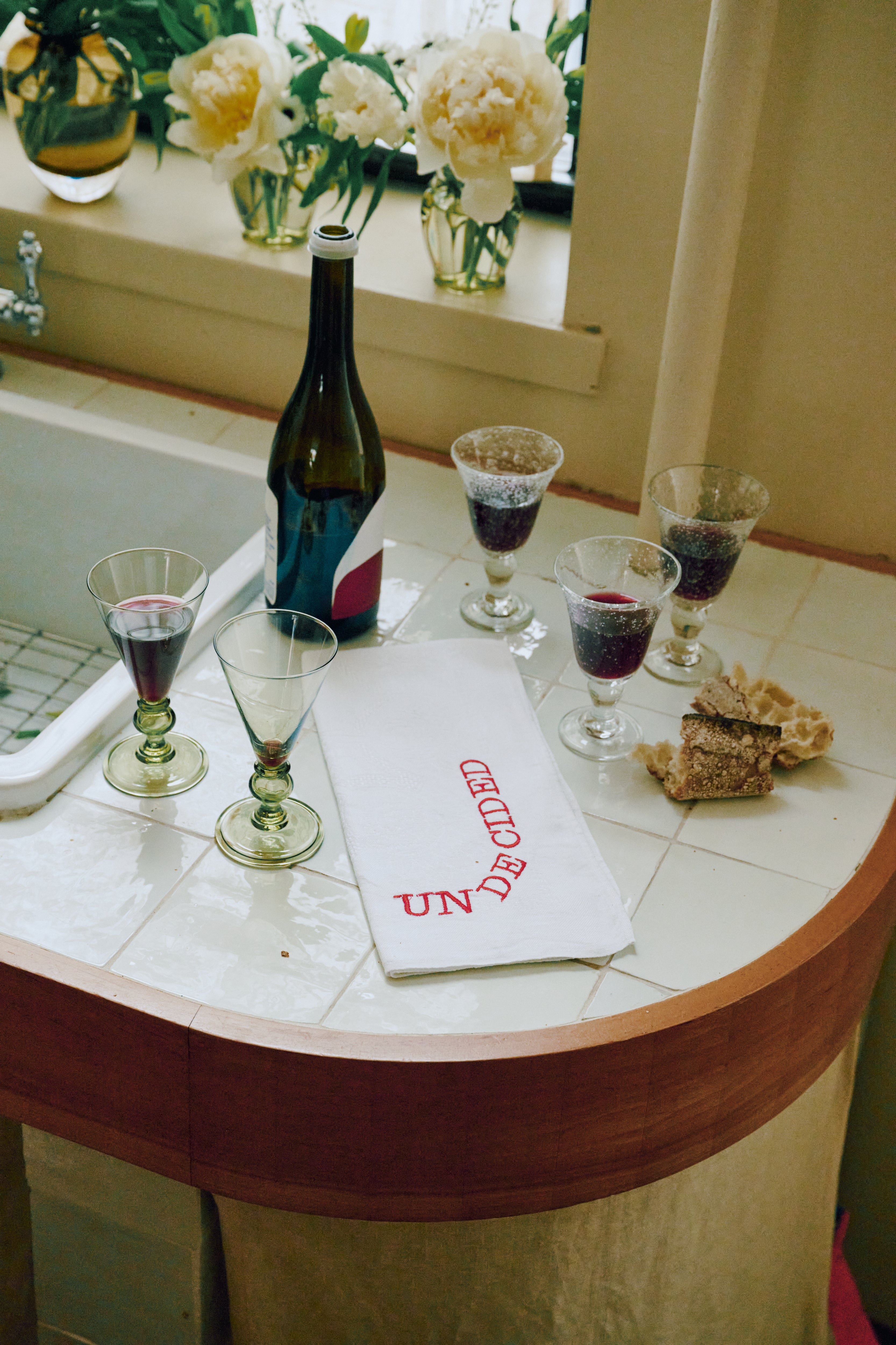 A bottle of red wine, four filled glasses, pieces of bread, and a Relationship Status Napkin from the set of 6 rest on a tiled countertop by a window with flowers.