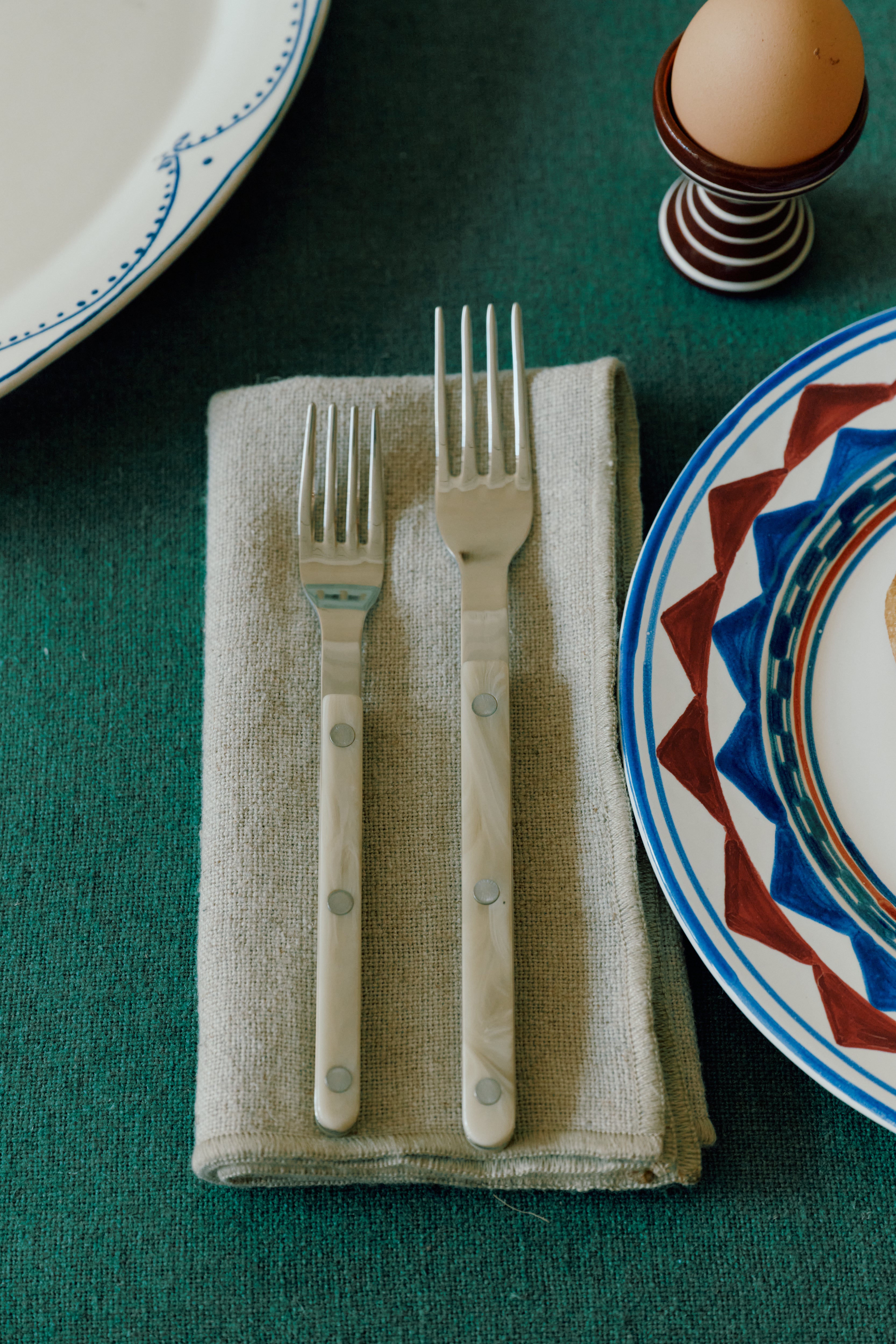 The Bistrot 5 Piece Cutlery Set features two forks resting on a folded napkin, with patterned plates and an egg cup on a green tablecloth for a Parisian bistro-inspired look.