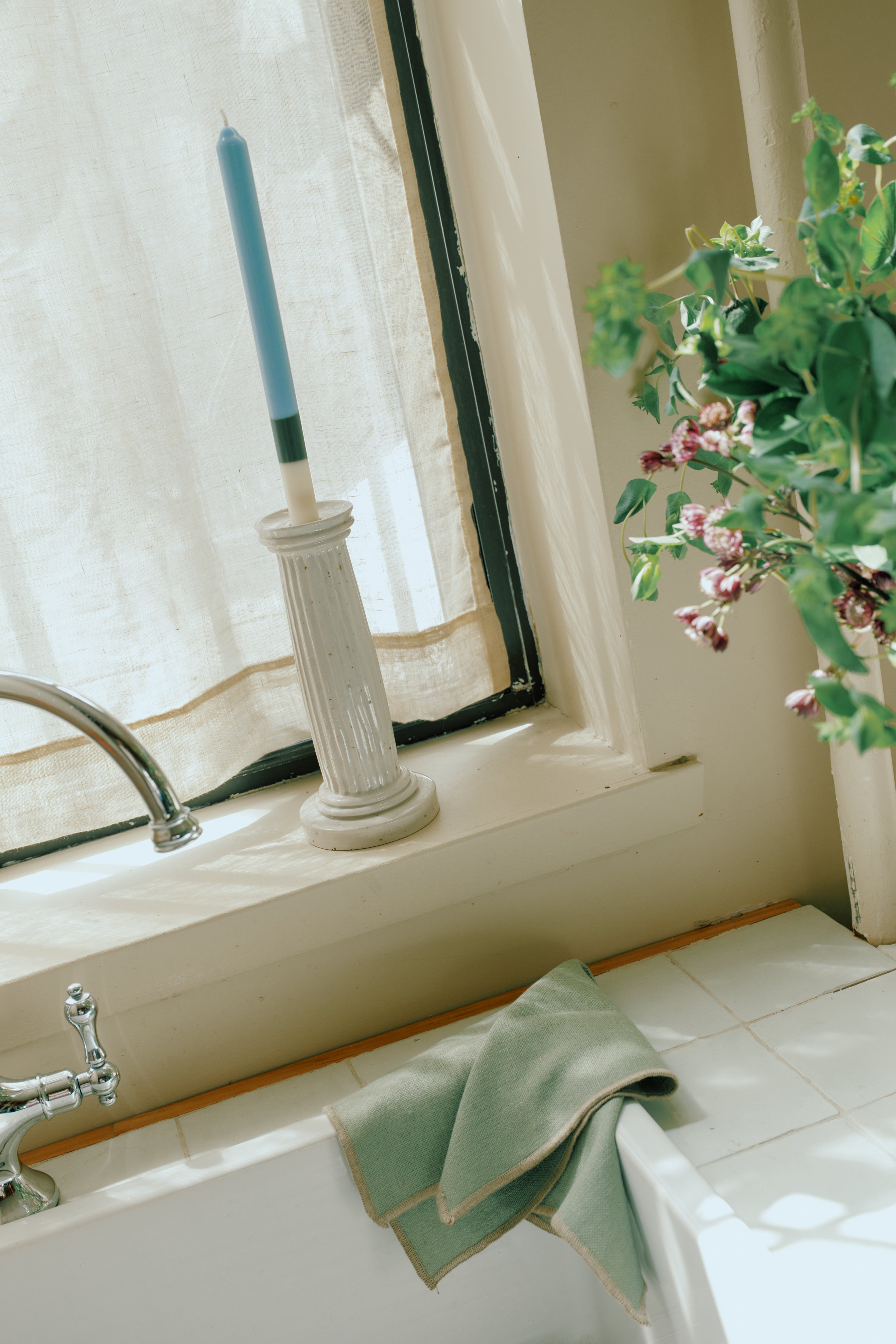 A blue candle in a white holder rests on the kitchen windowsill, with green dish towels, eco-friendly linen placemats and a set of 6 Corte Napkins, plus a nearby vase of flowers.