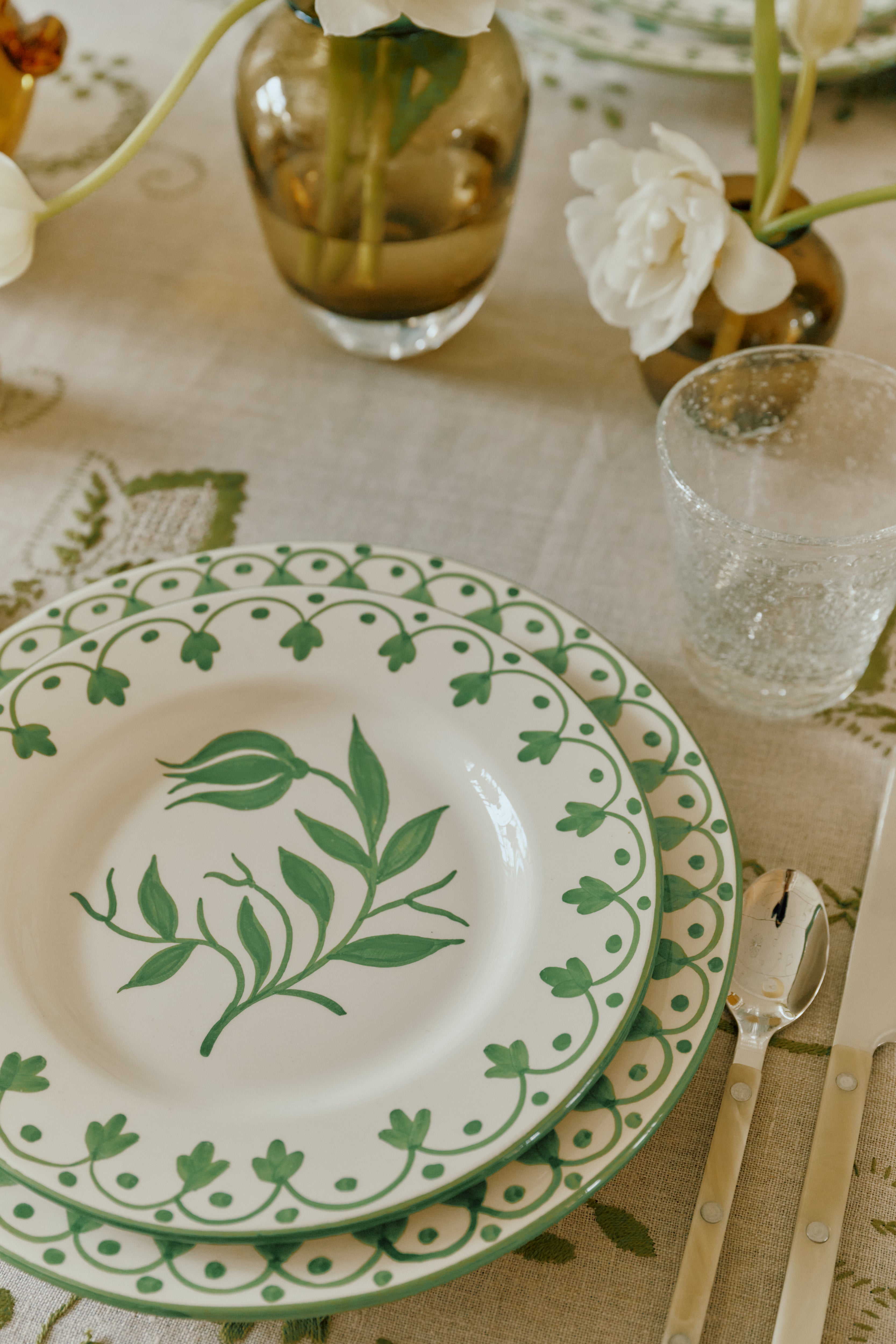 A table setting with two stacked Esmeralda Dinner Plates featuring green floral designs, a clear textured glass, knife, and fork on a patterned tablecloth with flowers in vases.