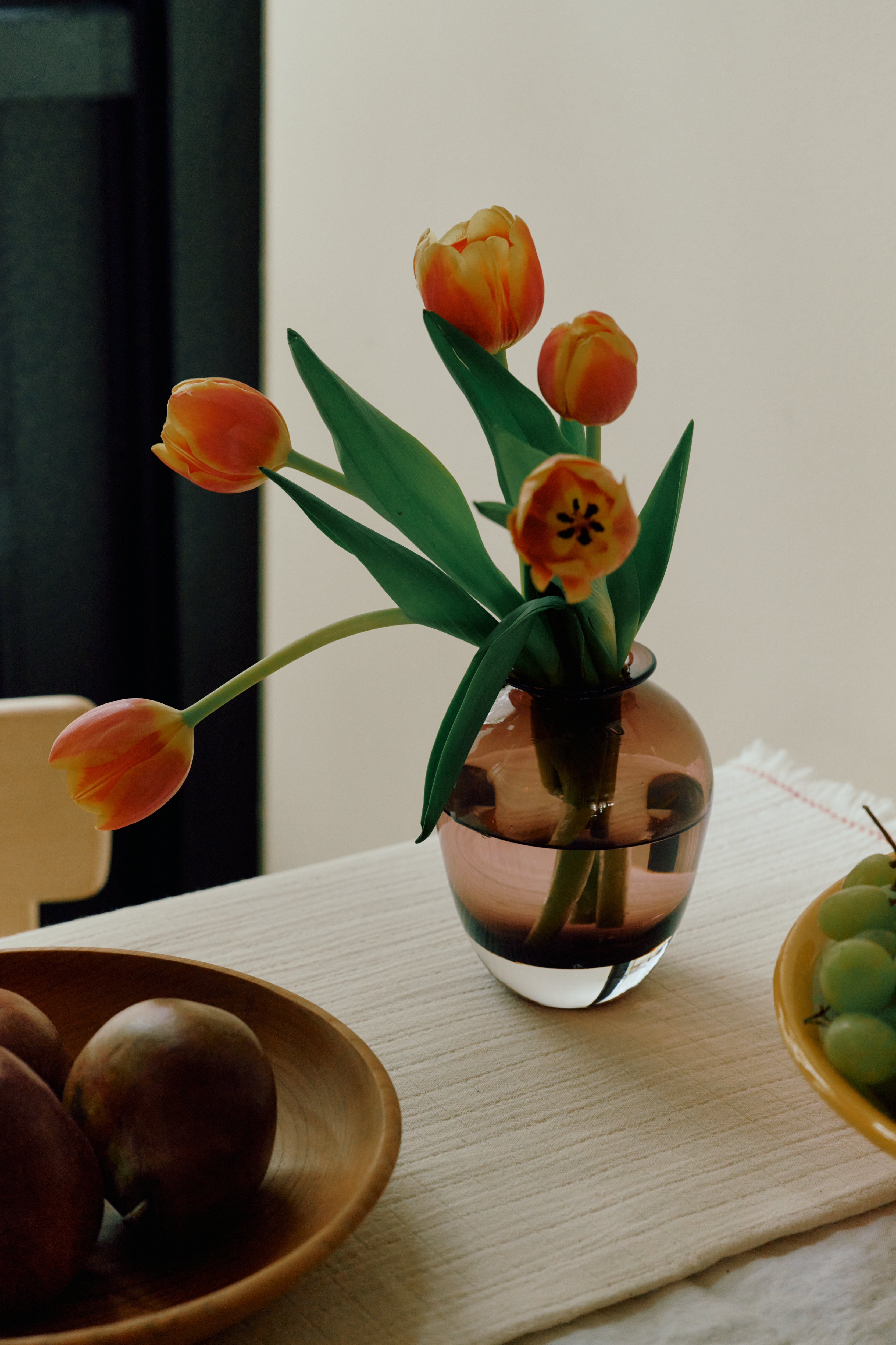The Gem Bud Vase, hand blown and small, holds orange tulips on a table beside a wooden fruit bowl and a plate of green grapes.