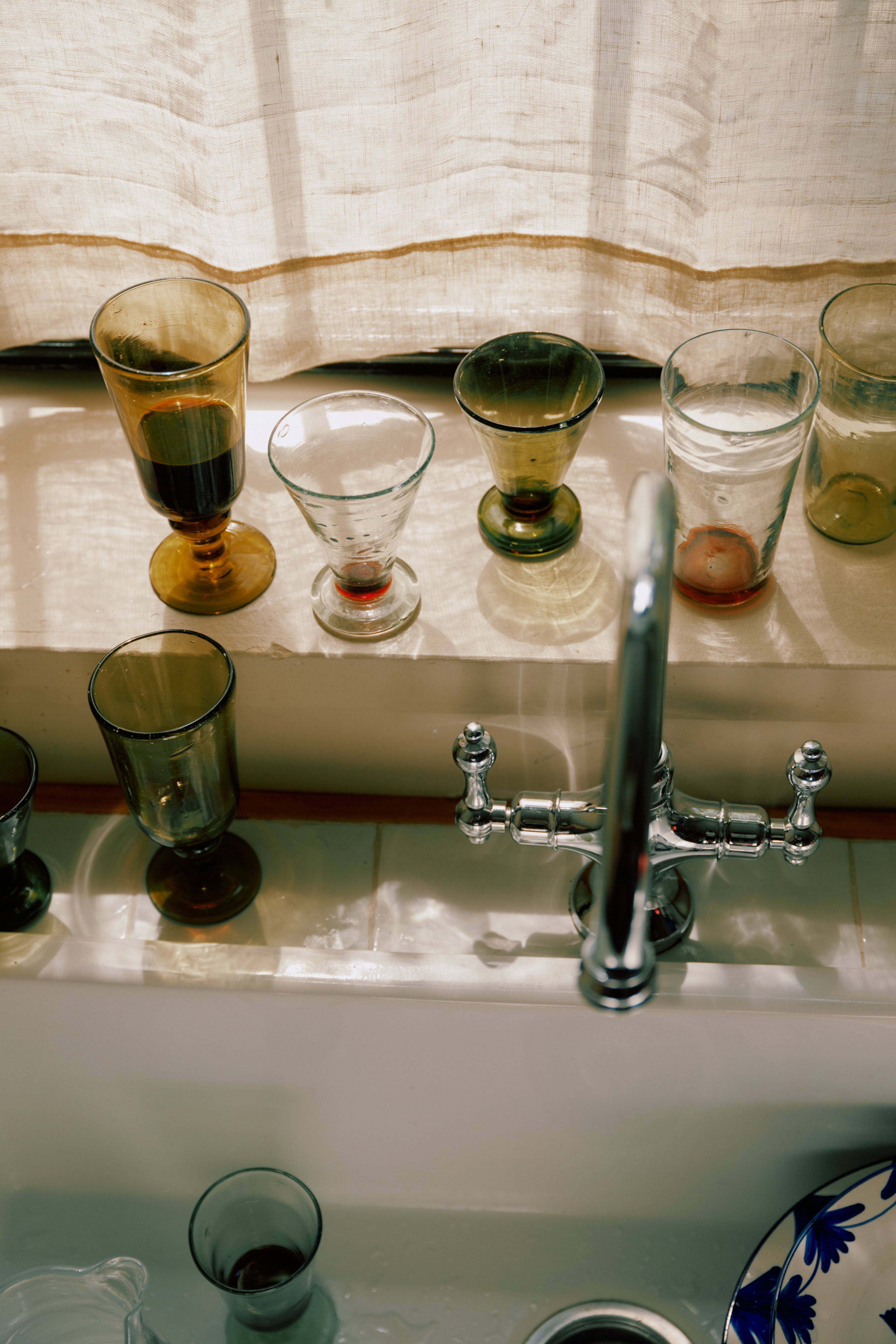 Several empty and partially filled Elodie Stemmed Wine Glasses (set of 6) are lined up on a windowsill above a kitchen sink with a silver faucet, sunlight streaming through a light-colored curtain.