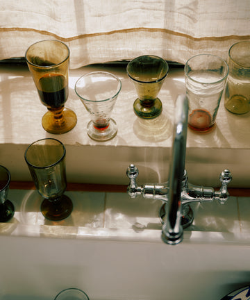 Several empty and partially filled Elodie Stemmed Wine Glasses (set of 6) are lined up on a windowsill above a kitchen sink with a silver faucet, sunlight streaming through a light-colored curtain.