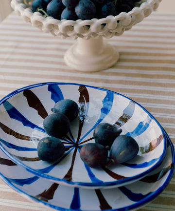 On a striped tablecloth, the Rayas Oval Serving Platter holds several dark purple figs, while a ceramic pedestal bowl filled with grapes sits behind it.