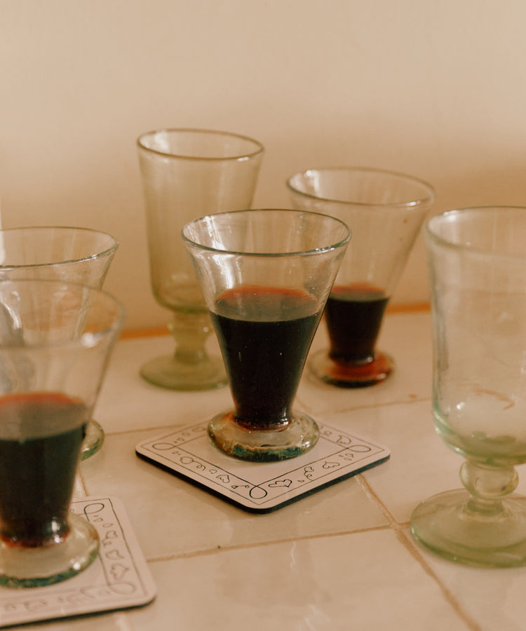 Six Elodie Footed Wine Glasses (set of 6), some filled with a dark liquid, are arranged on tiled surfaces, with two coasters visible beneath two of the recycled glass tumblers.
