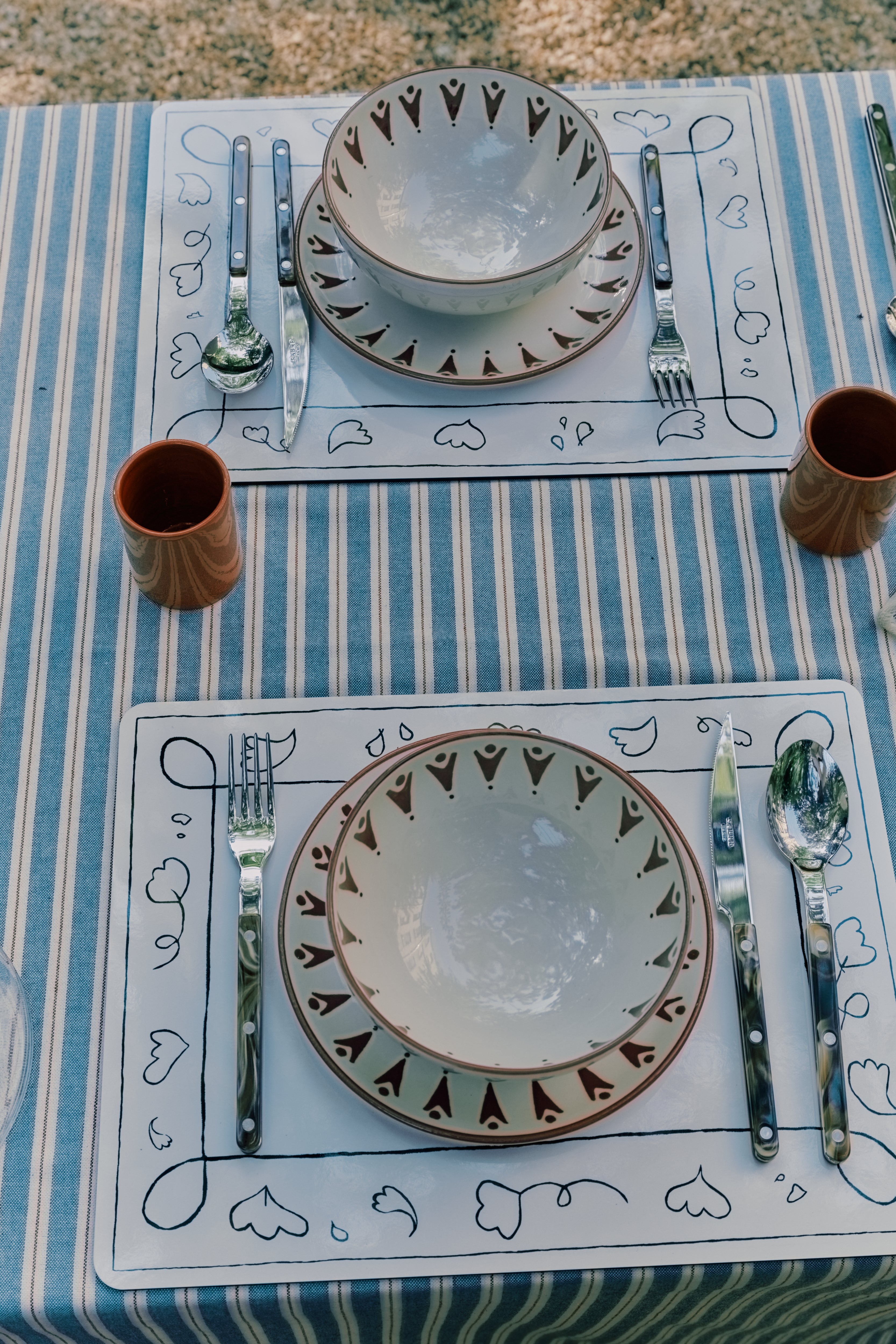 Two place settings on a striped tablecloth with patterned plates, bowls, silverware, and brown cups arranged neatly on wipeable Capriccio Placemats, Set of 6—featuring original decor inspired by PORTA artwork.