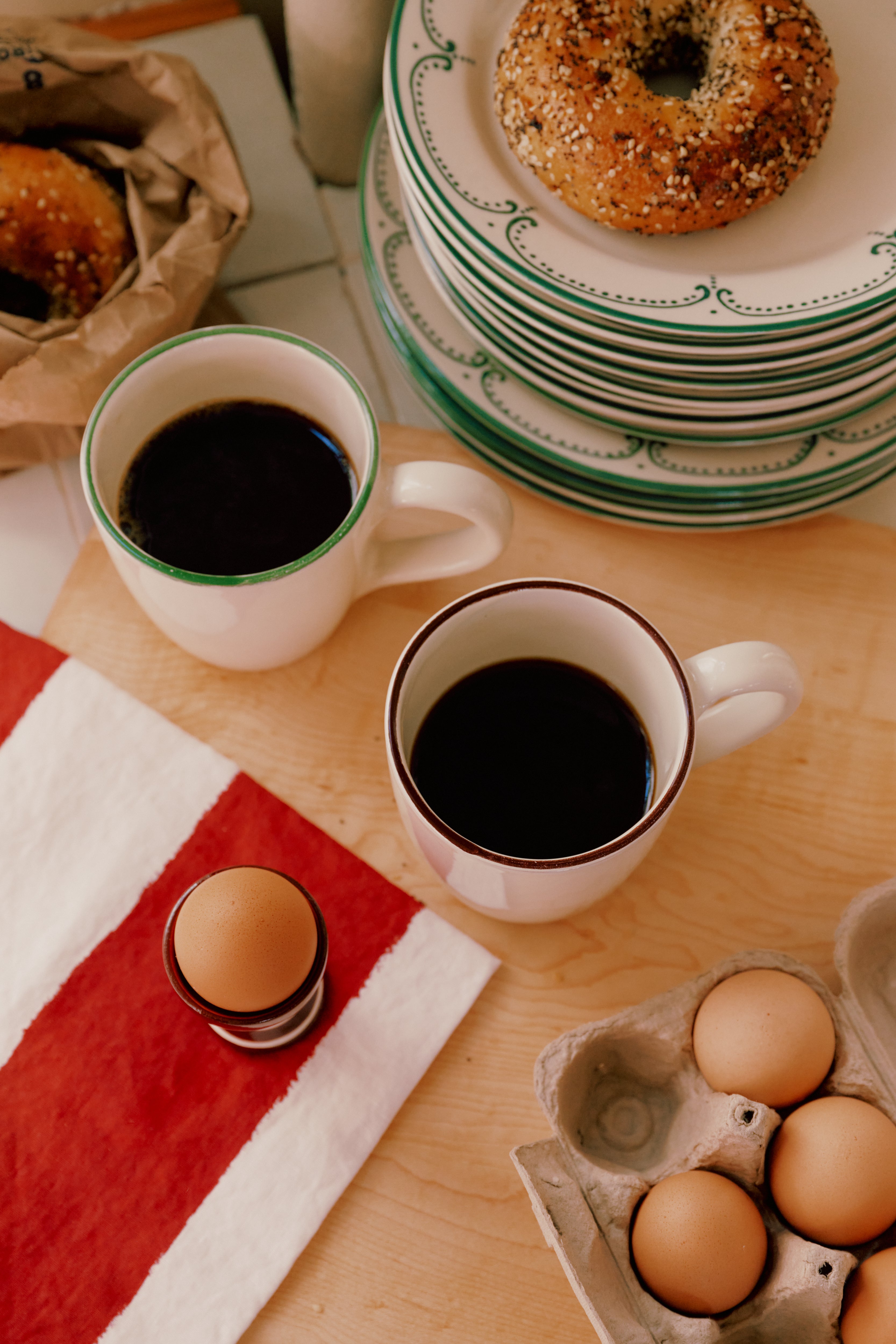Two Guido Mugs, a soft-boiled egg in a holder, a carton of eggs, a striped napkin, and bagels on plates are set on a wooden table.