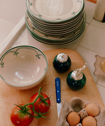 A stack of Arco Dinner Plate, Set of 4, and a bowl rest on a counter beside a cutting board holding tomatoes, a lemon wedge, eggs, a knife, and salt and pepper shakers.