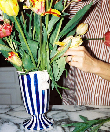 A person in a striped shirt arranges colorful tulips in a Thallo Pedestal Vase on a marble countertop.