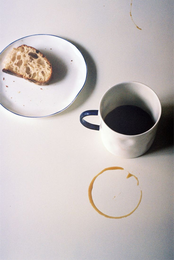 A Tea Mug filled with dark coffee sits beside a half-eaten slice of bread on a plate atop a table dotted with crumbs and coffee rings, showcasing minimalist design.