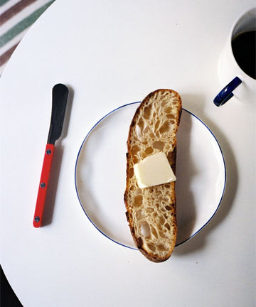 A slice of bread with butter rests on a plate beside a red-handled knife and black coffee, all atop a white table—perfectly complemented by tea served on our Cake Plate, Set of 4.