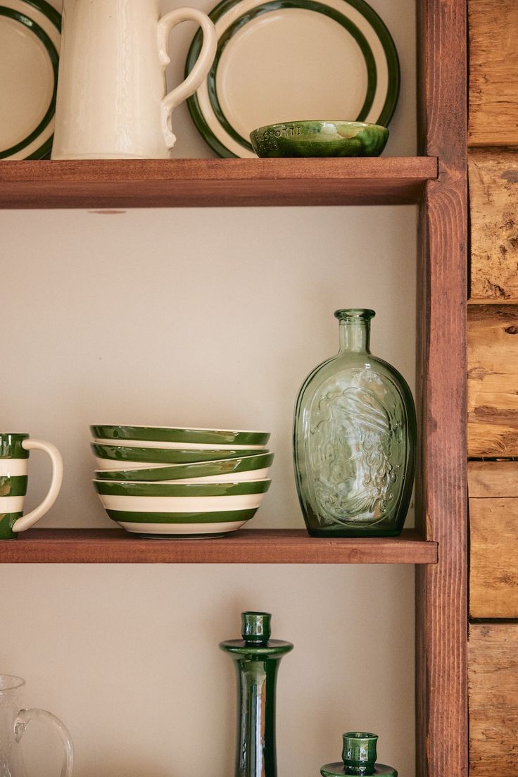 A wooden shelf showcases the Betty Cereal Bowl, green and white striped plates, a mug, a clear green glass bottle, and green glassware against a cream wall—an ideal setup for your breakfast collection.