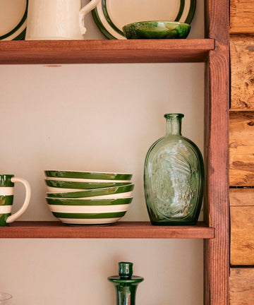 A wooden shelf showcases the Betty Cereal Bowl, green and white striped plates, a mug, a clear green glass bottle, and green glassware against a cream wall—an ideal setup for your breakfast collection.