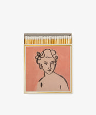 Luxury Matches come in an open matchbox with a pink cover, featuring a letterpress-printed line drawing of a person’s bust—curly hair, flowers, and a neutral expression—making them a unique keepsake gift.