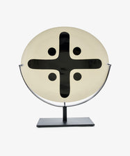 Motif 5, Oval is a round, cream-colored glazed earthenware piece featuring a bold black cross and four dots, displayed upright on a rectangular-based metal stand.