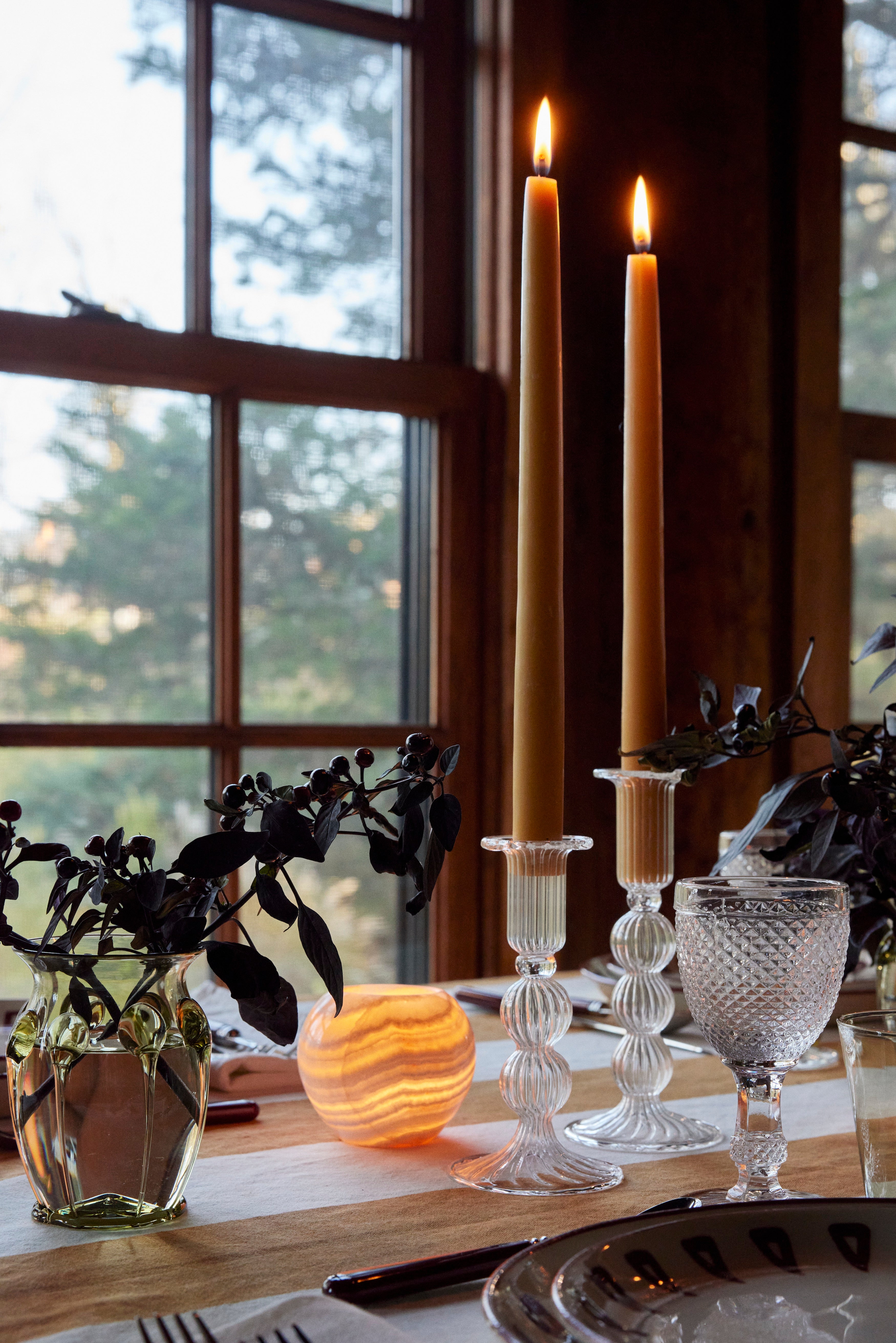 A table by a window features two lit tall candles in Akila Candleholders of Egyptian Alabaster, a water glass, dark foliage in a vase, and a glowing round lamp.