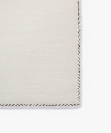 A close-up of the Filial Napkin, Set of 6, in cream with a grid pattern and dark branch stitching along the edge, displayed on a white tabletop background.