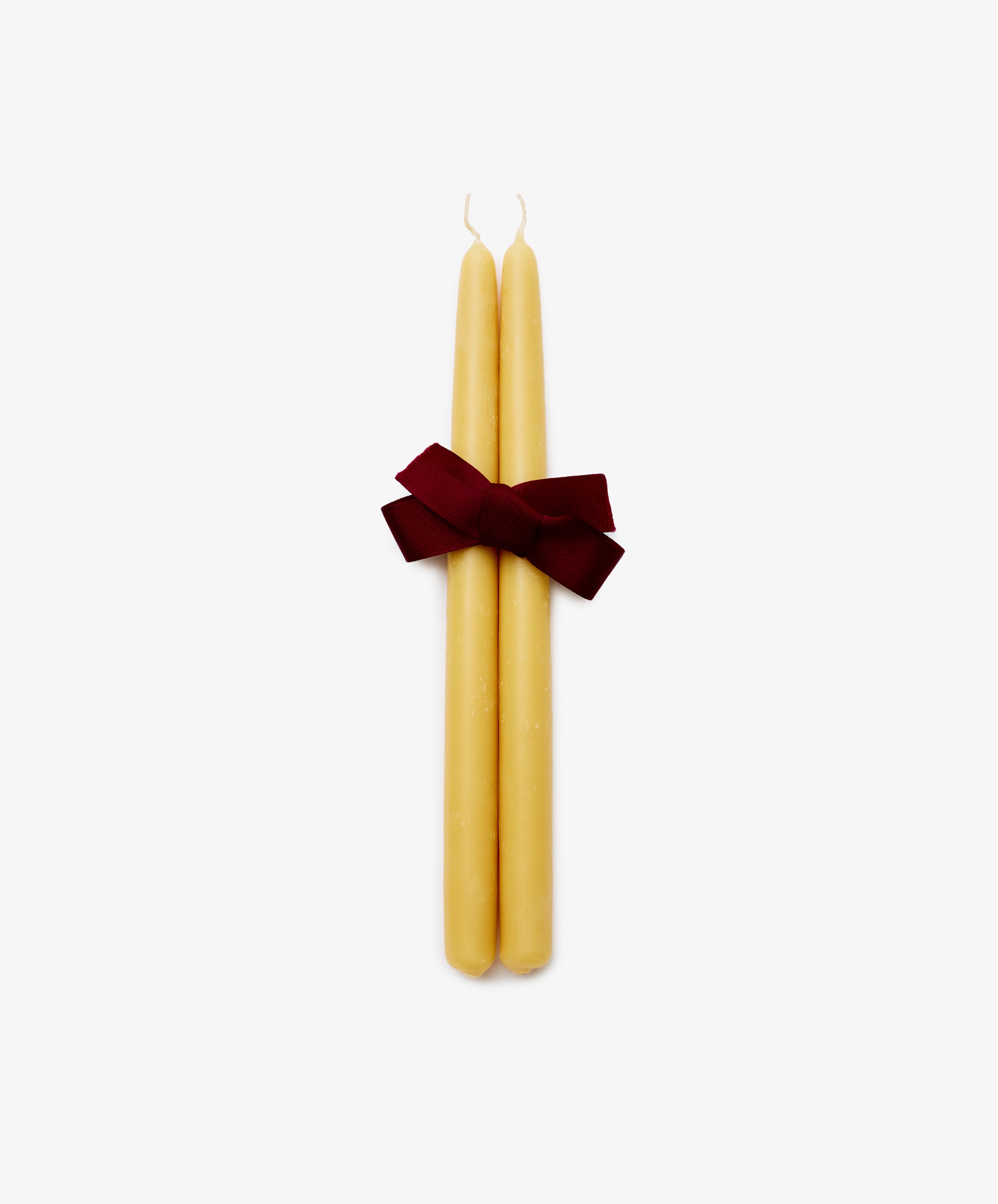 The Tapered Candles, Set of 2, in yellow are tied with a dark red ribbon bow and displayed on a plain white background. Each candle offers up to 12 hours of burn time.