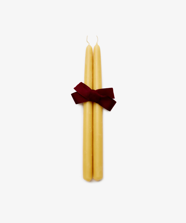 The Tapered Candles, Set of 2, in yellow are tied with a dark red ribbon bow and displayed on a plain white background. Each candle offers up to 12 hours of burn time.