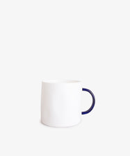 The Tea Mug features a minimalist plain white ceramic body with a dark blue handle, handcrafted quality, and is shown against a light gray background.