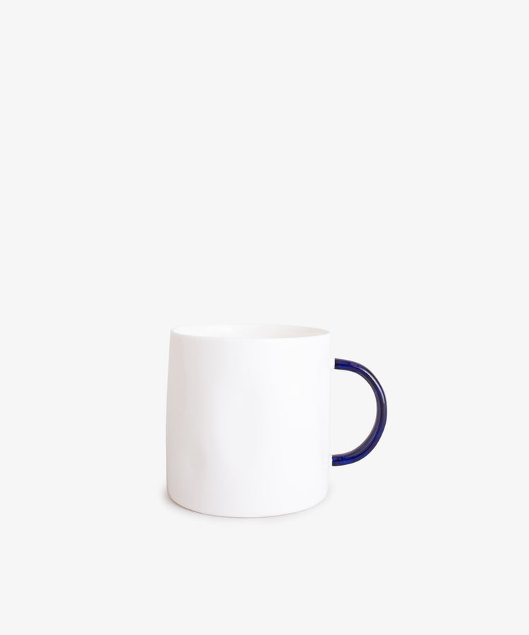 The Tea Mug features a minimalist plain white ceramic body with a dark blue handle, handcrafted quality, and is shown against a light gray background.