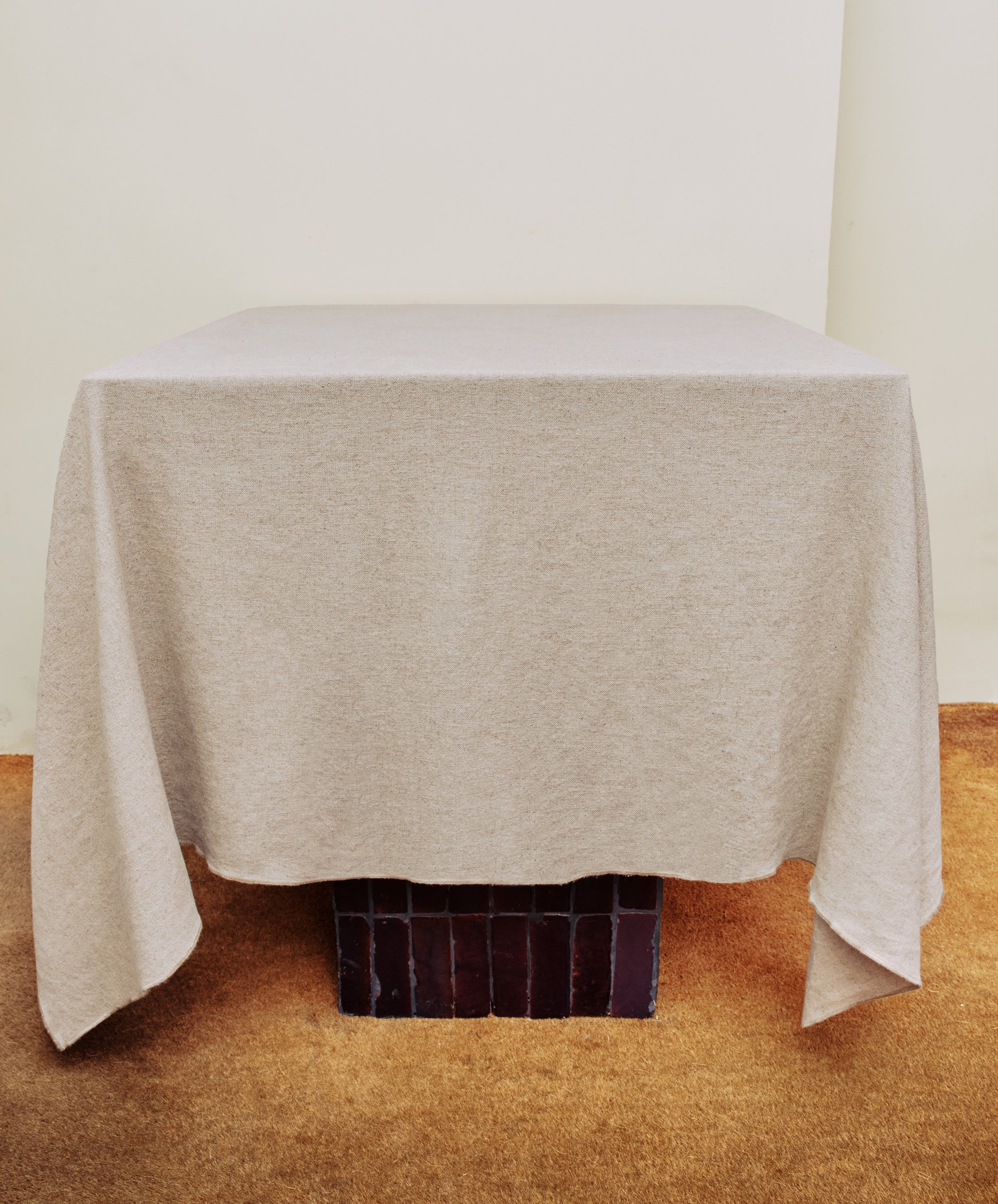 A square table with a Corte Tablecloth in plain beige stands on a brown carpeted floor against a cream-colored wall.