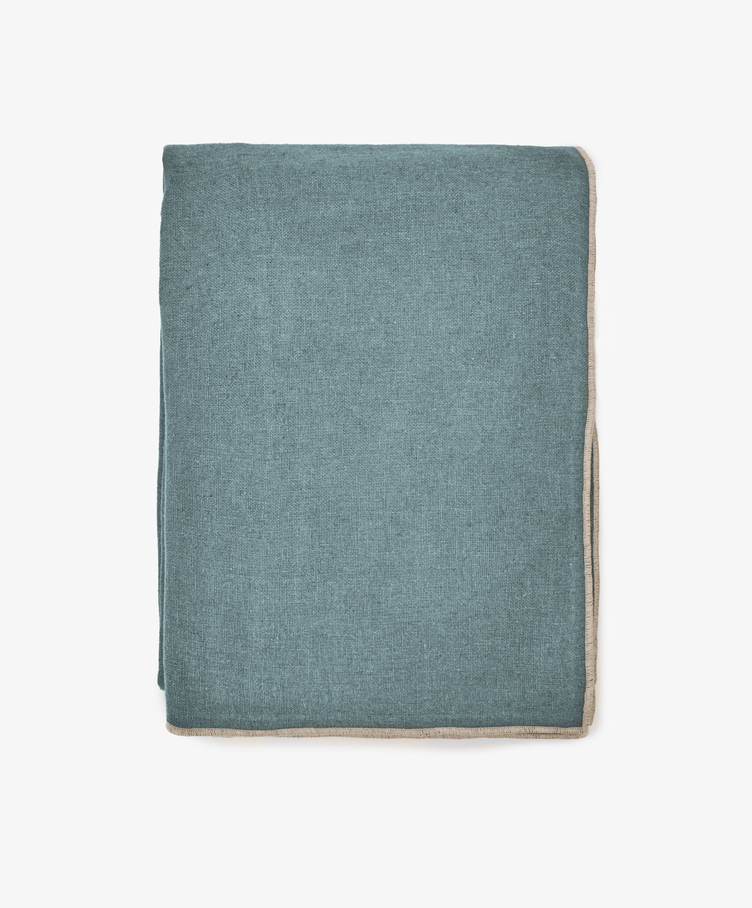 The Corte Tablecloth, a neatly folded rectangular piece in teal with beige trim, exudes sophistication as it is displayed against a plain white background.