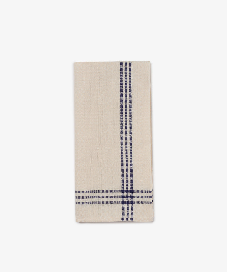 The Tilde Tea Towel, made from a beige cotton-linen blend, features two navy blue plaid stripes running vertically and horizontally near the edge and is shown folded on a white background.