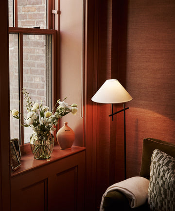 A cozy corner with a lit floor lamp, green sofa, patterned pillow, and a window ledge holding a vase of white flowers and the Hugo Money Pot. Brick wall visible through the window.