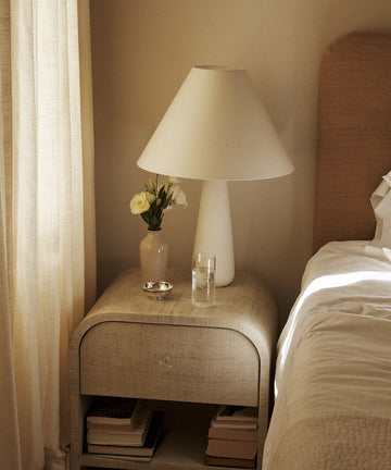 A bedside table next to a bed with white linens holds a lamp, a vase of white flowers, the Deco Tall Water Glass, Set of 6 in cobalt blue, and a small dish, with books stacked on the lower shelf.