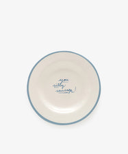 The You silly sausage! plate is a white ceramic dish, handmade in Portugal by artist Laetitia Rouget, featuring a blue rim and the phrase “you silly sausage!” handwritten in blue at the center.