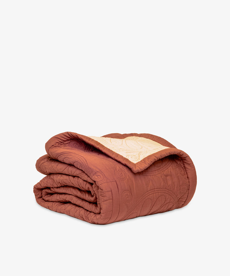 The Dawn Twin Bedspread, a folded, quilted blanket with a rust-colored exterior and beige interior that highlights Swedish craftsmanship, is displayed against a plain white background.