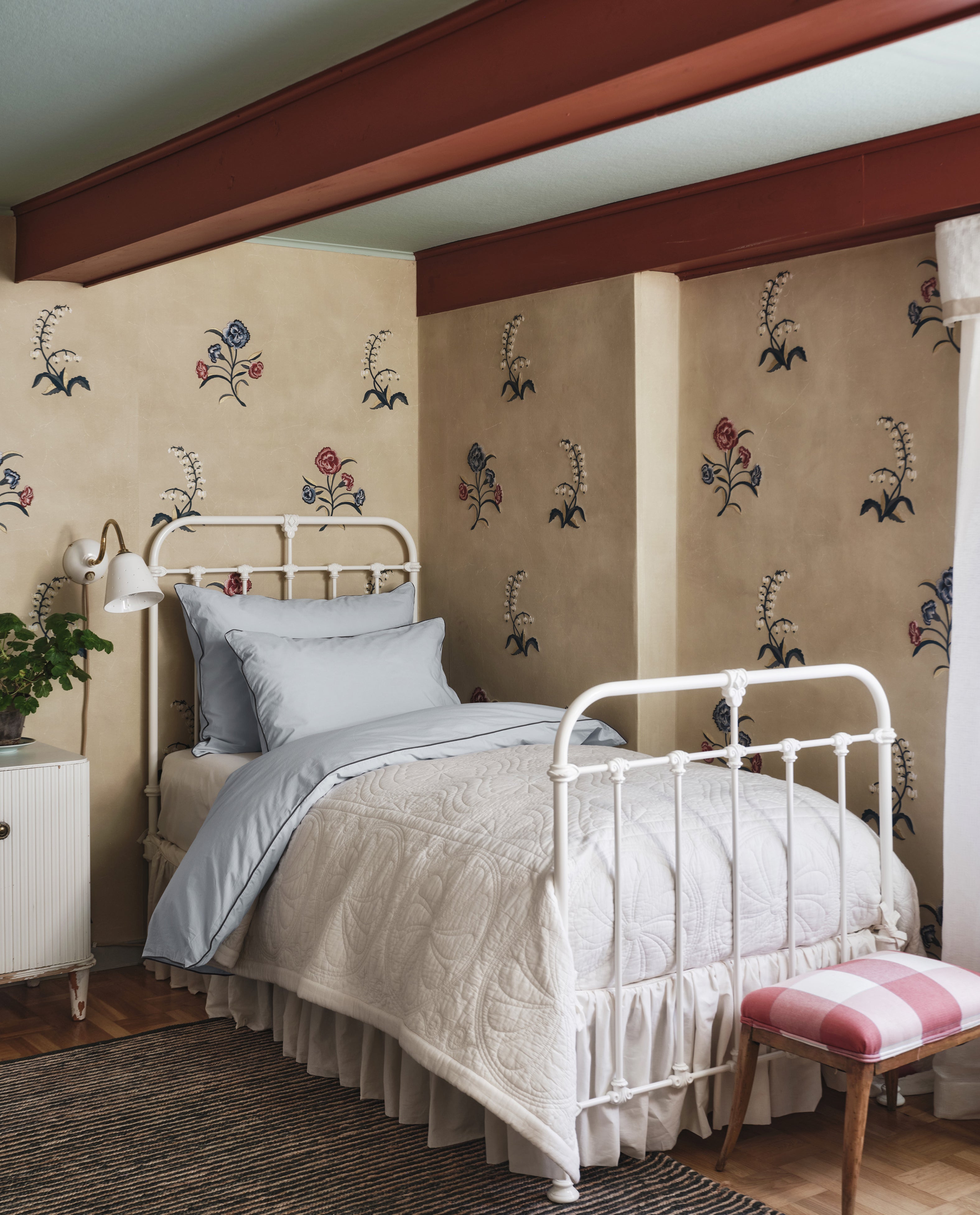 A Dawn Twin Bedspread in light blue covers a neatly made white metal bed set against a beige floral-patterned wall in a cozy bedroom with exposed red beams.
