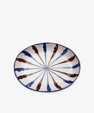 The Rayas Oval Serving Platter is a handmade ceramic dish with a vibrant blue and brown radial striped pattern on a white background.