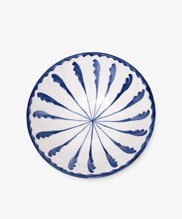 The Rayas Serving Bowl is a round ceramic piece, hand-painted with a blue leaf-like radial pattern on a white base, highlighting its unique handmade charm.