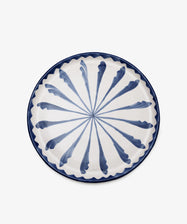 The Rayas Serving Plate is a hand-painted ceramic piece with a vibrant blue radial leaf pattern on a white base and a blue wavy border, shown against a plain white background.
