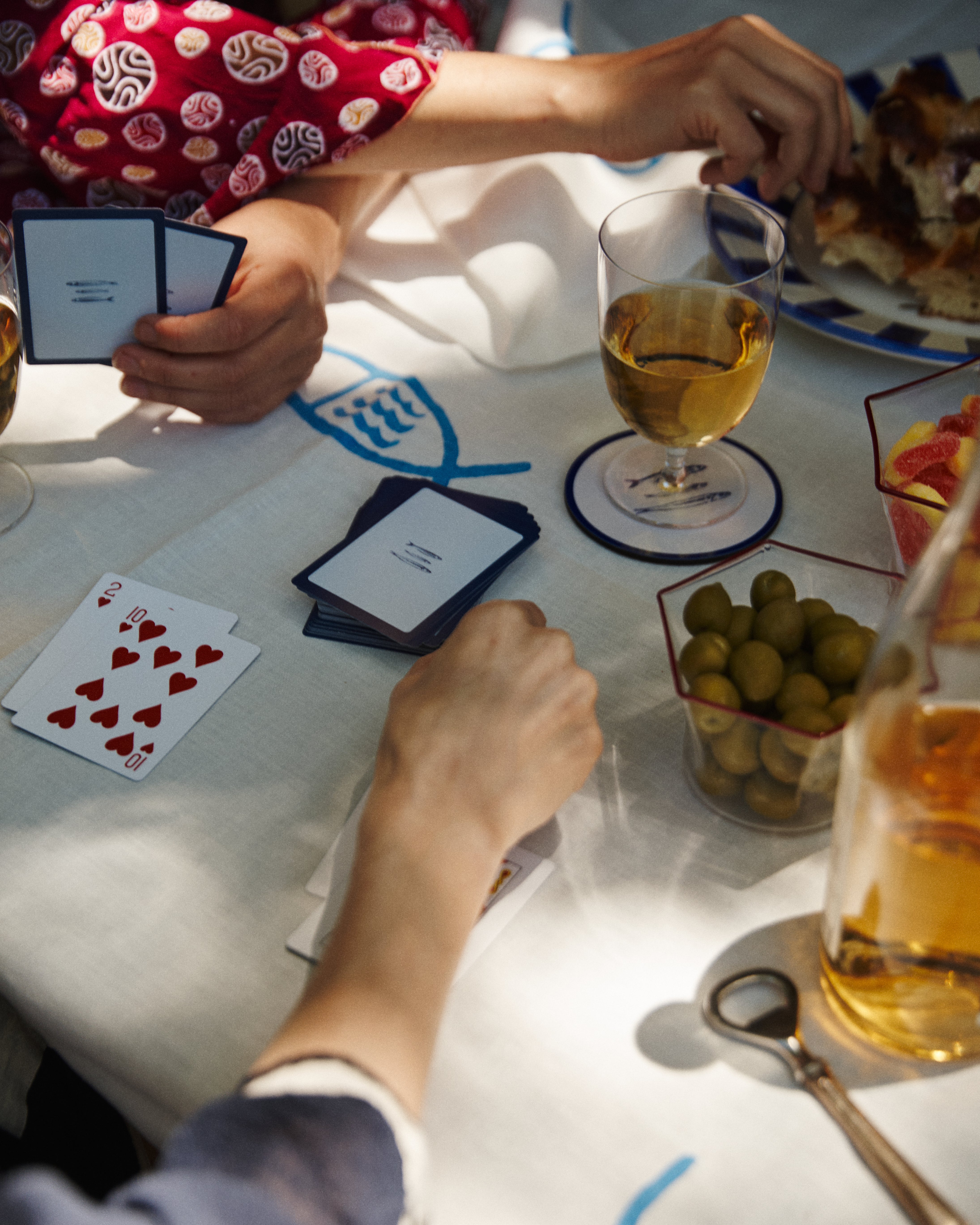 Three people enjoy a lively evening playing cards at a table with wine, olives, snacks, and food on a white-and-blue cloth, using PORTA Playing Cards.